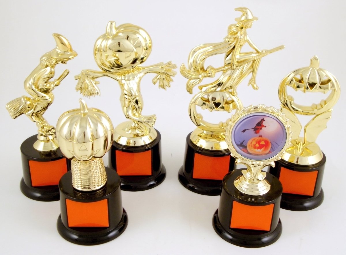 Six gold-toned Halloween-themed trophy figures with black bases and orange square panels, arranged on a white studio background. Designs include a witch riding a broom, a scarecrow, a flying witch, a jack-o'-lantern, a pumpkin, and a circular pumpkin-and-witch emblem.
