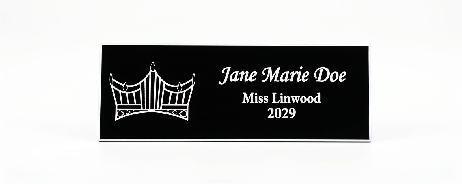 A rectangular black Name Badge  with white engraved text reading 'Jane Marie Doe, Miss Linwood, 2029' and a white crown graphic on the left, shown on a plain white background.