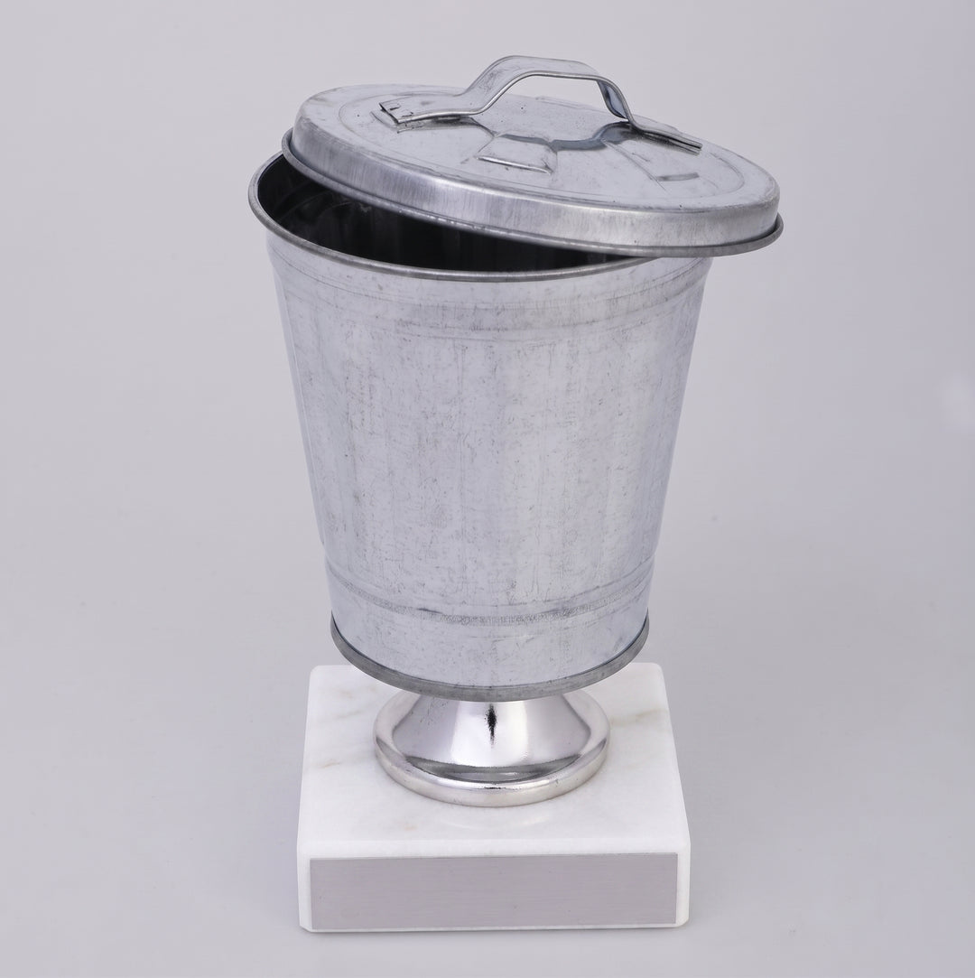 Galvanized metal wastebasket Trophy on a polished silver pedestal base with a white marble base, photographed against a neutral light gray background.