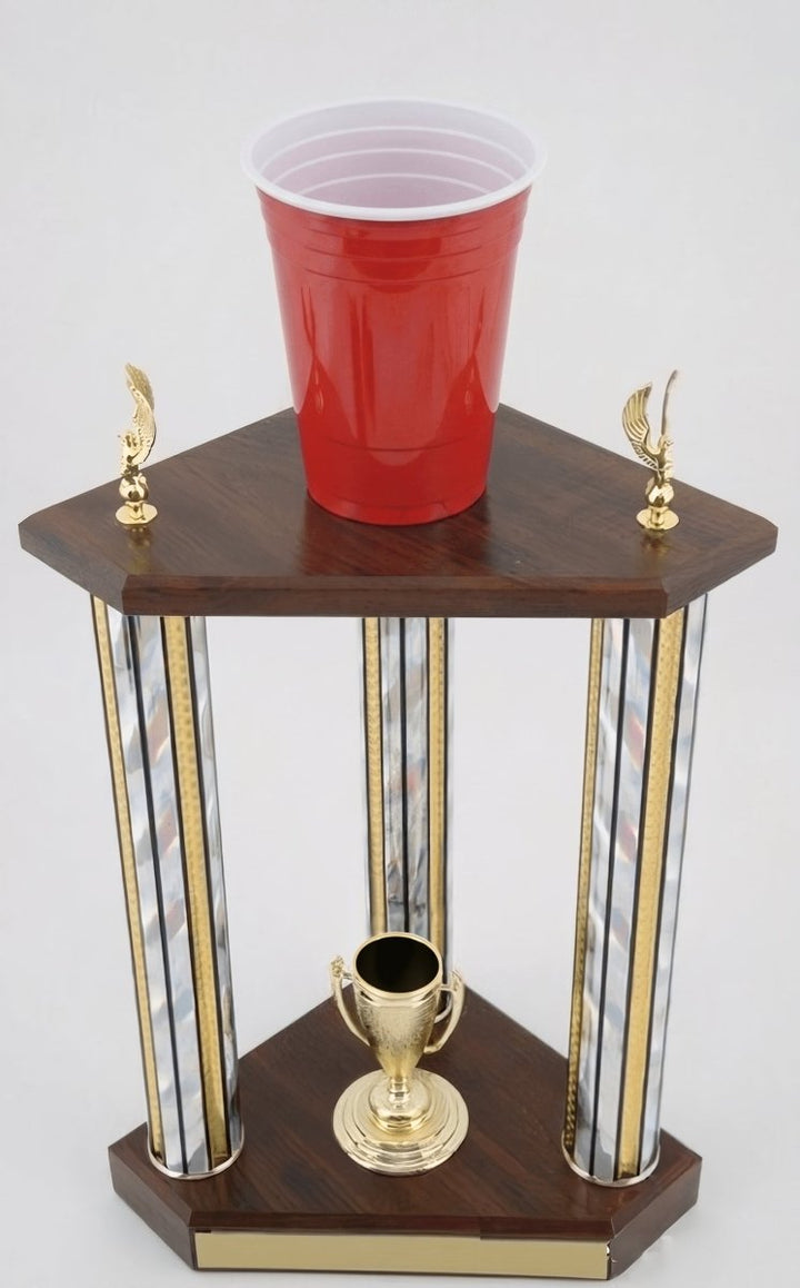 A triangular styled wooden trophy stand with gold and silver striped columns, a small gold cup on the bottom shelf, and a red plastic cup resting on the top platform, photographed against a light gray background.