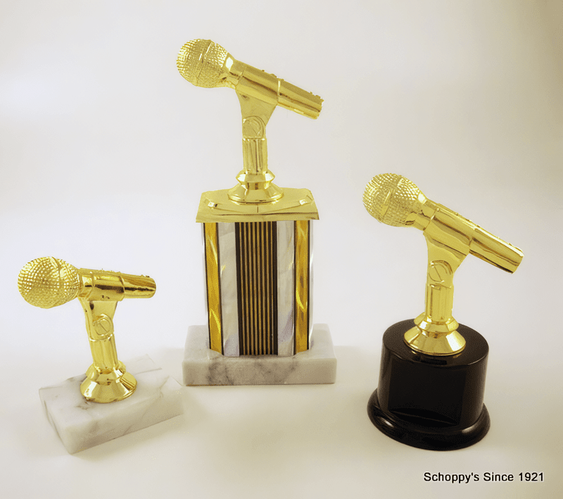 Three gold microphone-shaped trophy stands displayed on white and black bases against a clean white background. The center stand features a striped gold, white, and black column, while the left stand sits on a white marble base and the right stand has a glossy black cylindrical base.