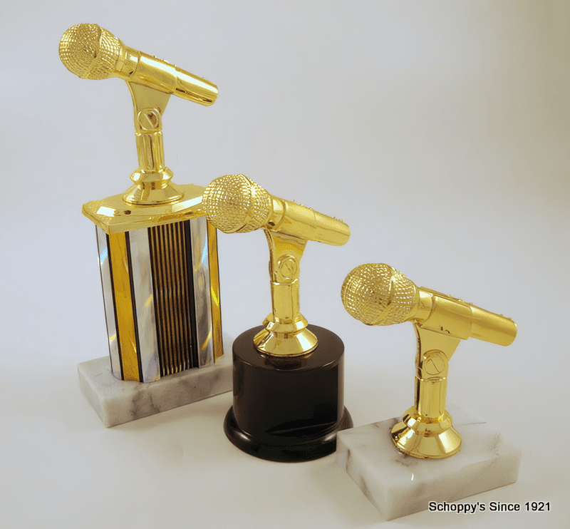 Three microphone-shaped trophy awards displayed on marble and dark bases against a light gray background. The trophies feature shiny gold microphone heads and stems, with one on a silver striped column, one on a black cylinder, and one on a white marble base.