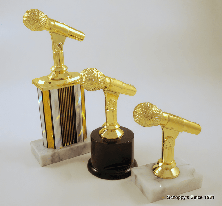 Three microphone-shaped trophy awards displayed on marble and dark bases against a light gray background. The trophies feature shiny gold microphone heads and stems, with one on a silver striped column, one on a black cylinder, and one on a white marble base.